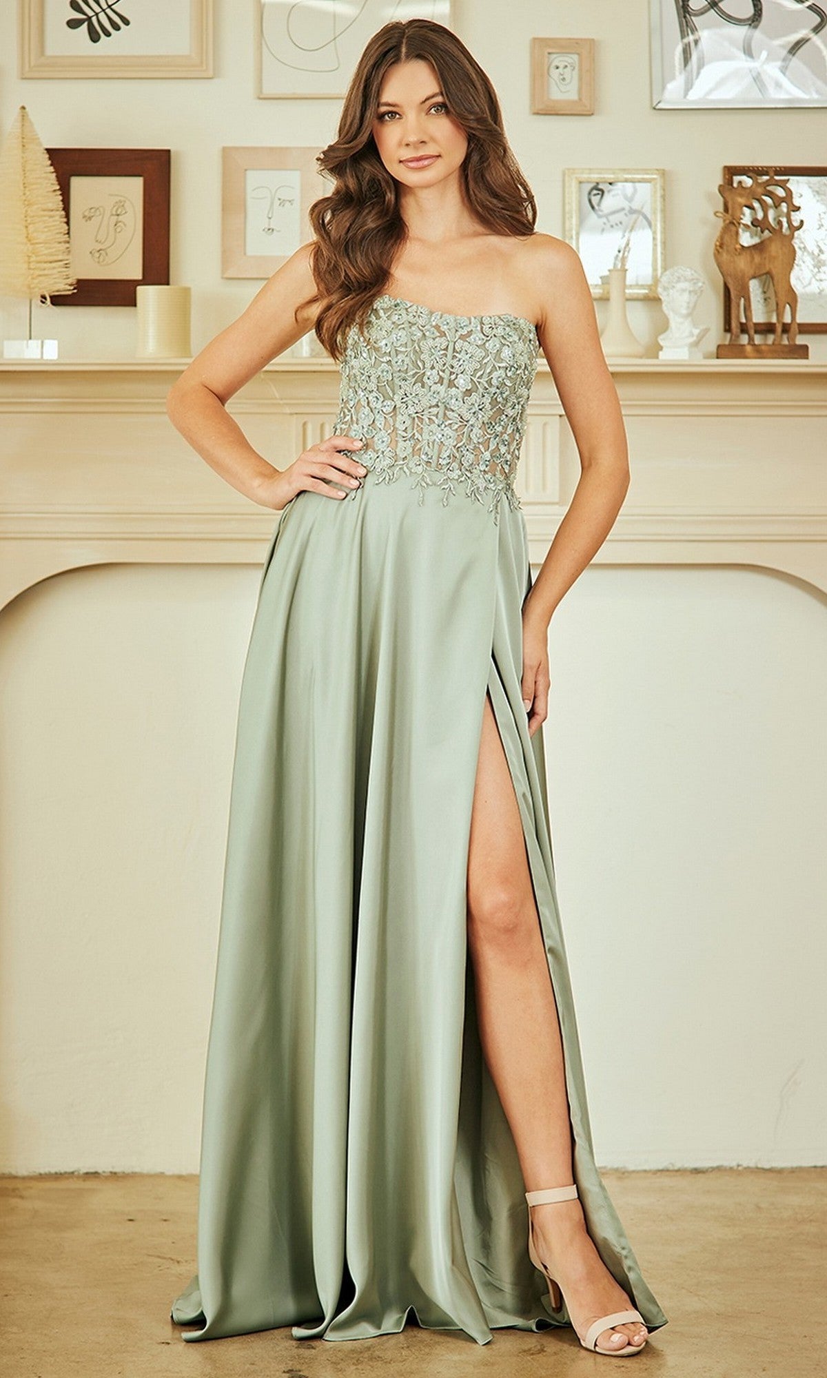 A woman in the Amelia Couture 6204 long prom dress, featuring a pale green A-line silhouette, beaded bodice, and thigh-high slit, stands indoors before a decorated mantel.